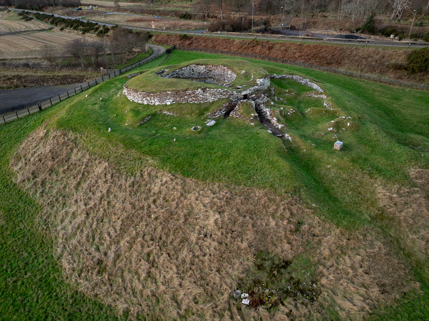 A stone broch on a grass mound, there are roads in the background. 
