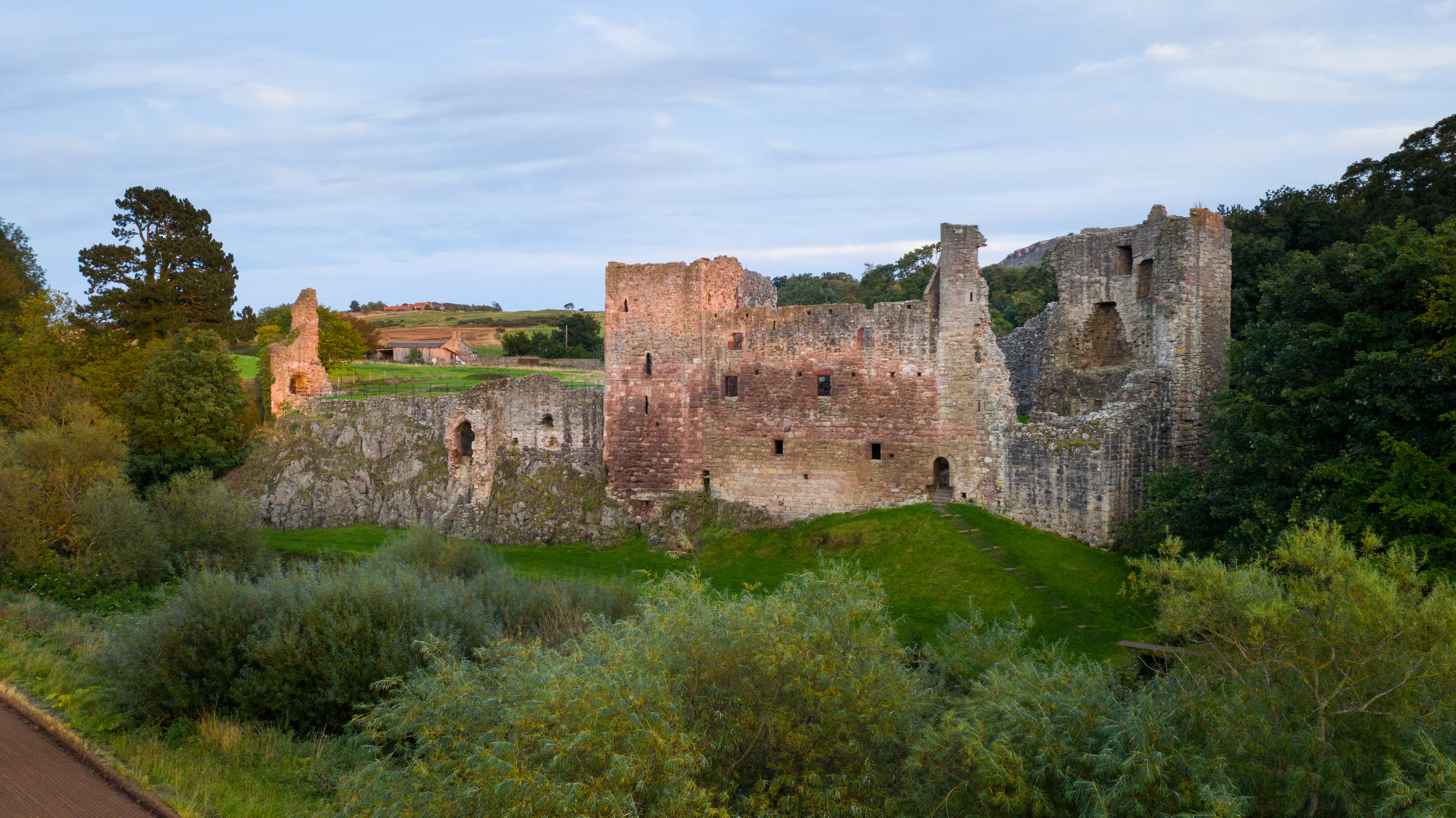 Wide view of Hailes Castle ruins with tall stone walls and towers, set against a backdrop of trees and open countryside under a pale blue sky.