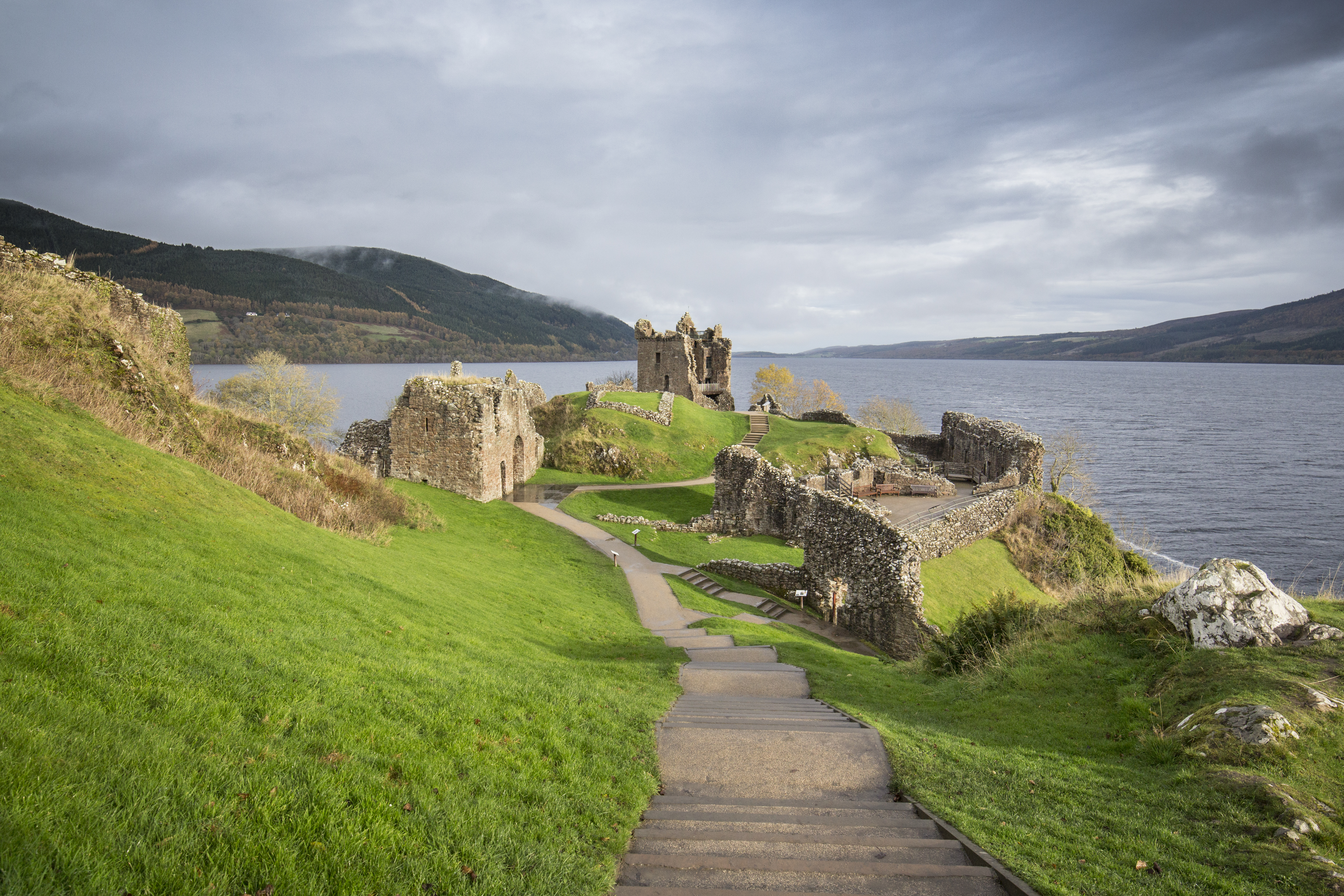 An empty path with steps leading through castle ruins towards a tower. There is a large loch in the background, 