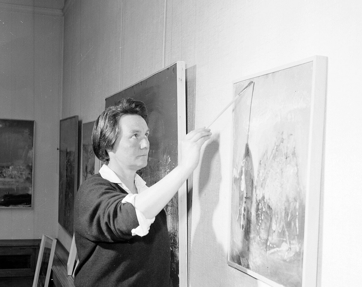 A woman holding a paintbrush and looking at her canvas.