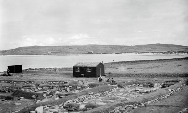 An excavation at Jarlshof, Shetlands. There is a small building in the middle of the photo, and two people in the middle working on the ground. You can see stones on the ground marking a settlement. There is a body of water in the background. 