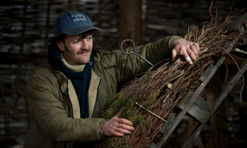 Man in cap with moustache fixes heather thatch onto a frame.