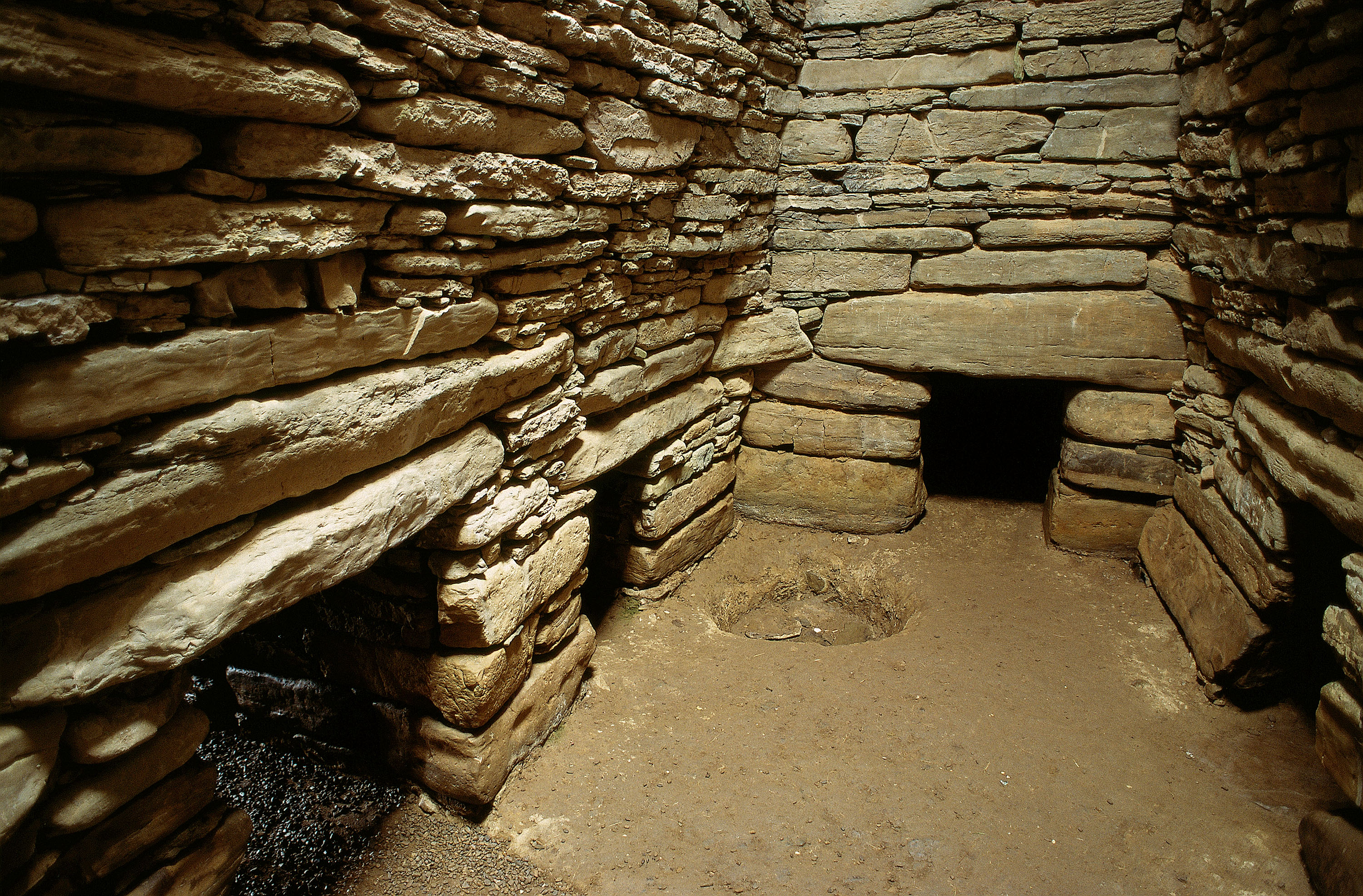 Interior chamber of Quoyness Chambered Cairn, showing thick drystone walls built from flat stacked stones. Several low side‑cells open off the main chamber, each with a small rectangular entrance framed by stone slabs. The floor is bare earth with a shallow circular depression in the centre. The stone walls curve inward slightly toward the roof, and the space is dimly lit, highlighting the textures of the ancient masonry.