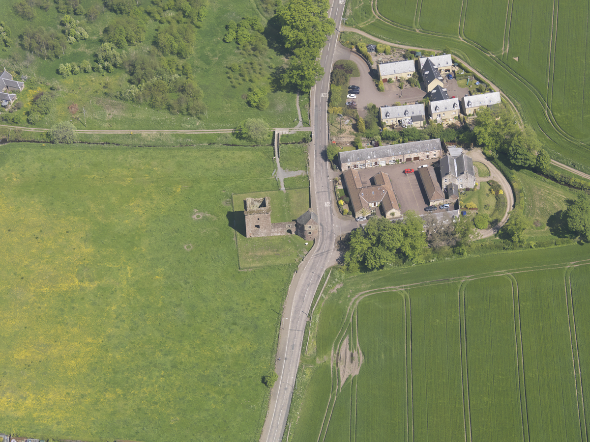 A high‑level aerial photograph showing Burleigh Castle standing alone in a large green field. The castle consists of two rectangular stone towers connected by low, ruined walls. A narrow road curves past the site, separating the field from a cluster of modern houses and farm buildings on the right side of the image. Surrounding the area are agricultural fields with visible tractor lines and patches of trees and hedgerows. The castle ruins appear small within the wide landscape.