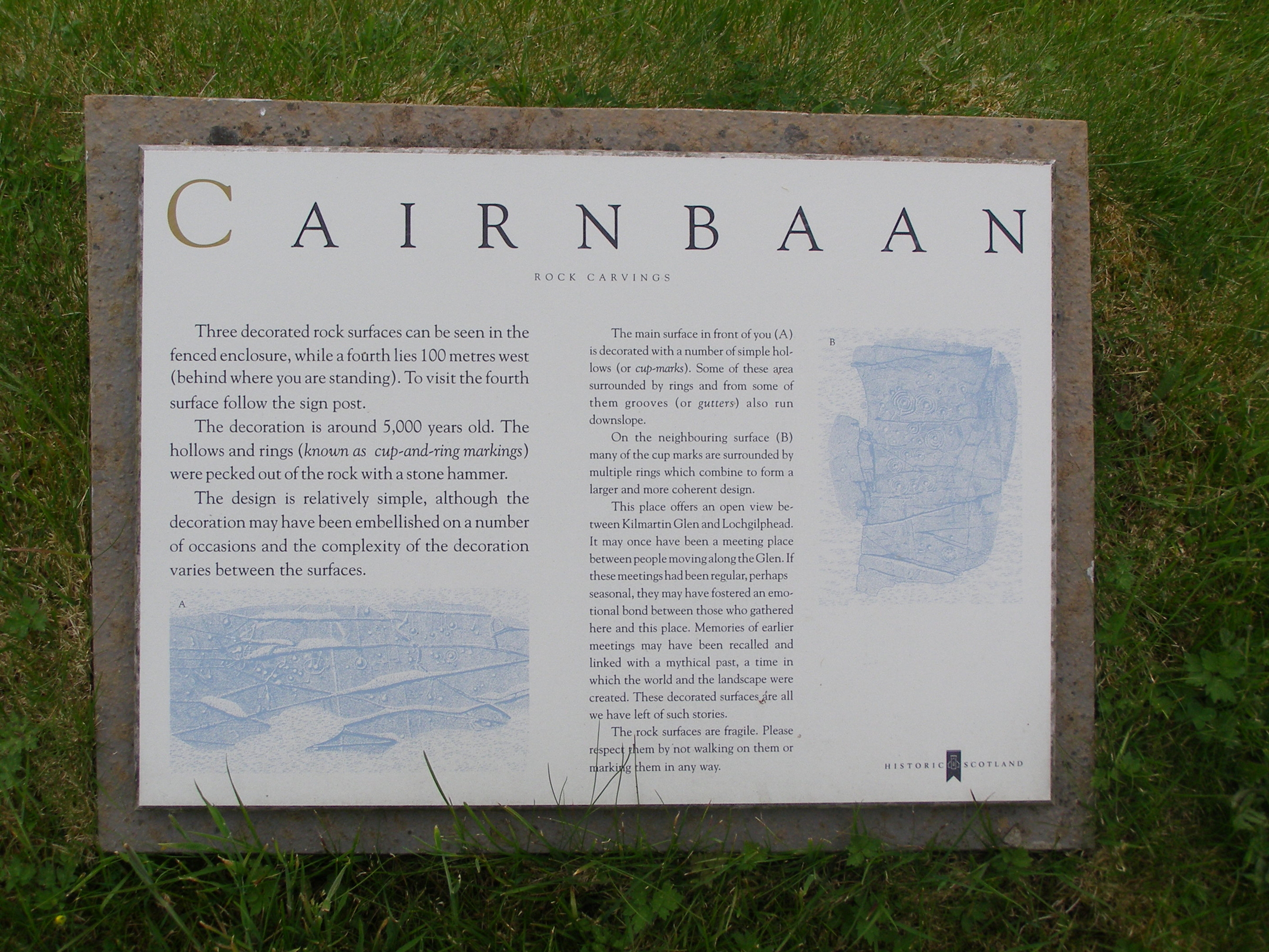 A rectangular outdoor information panel titled “Cairnbaan Rock Carvings,” mounted on a stone slab and set in grass. The panel contains explanatory text about the prehistoric rock art at Kilmartin Glen, along with faint blue illustrations of the carved rock surfaces.
