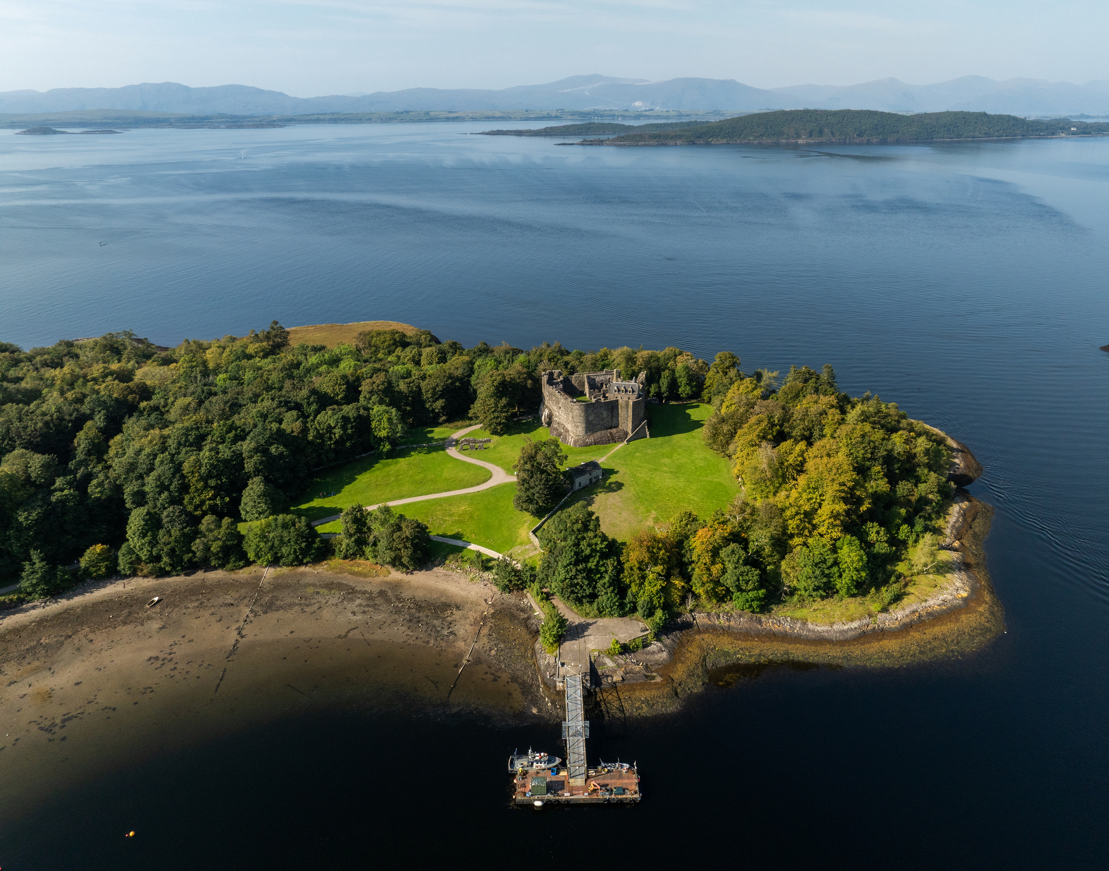 Aerial view of a castle on a grassy peninsula extending into a wide expanse of water with distant hills.