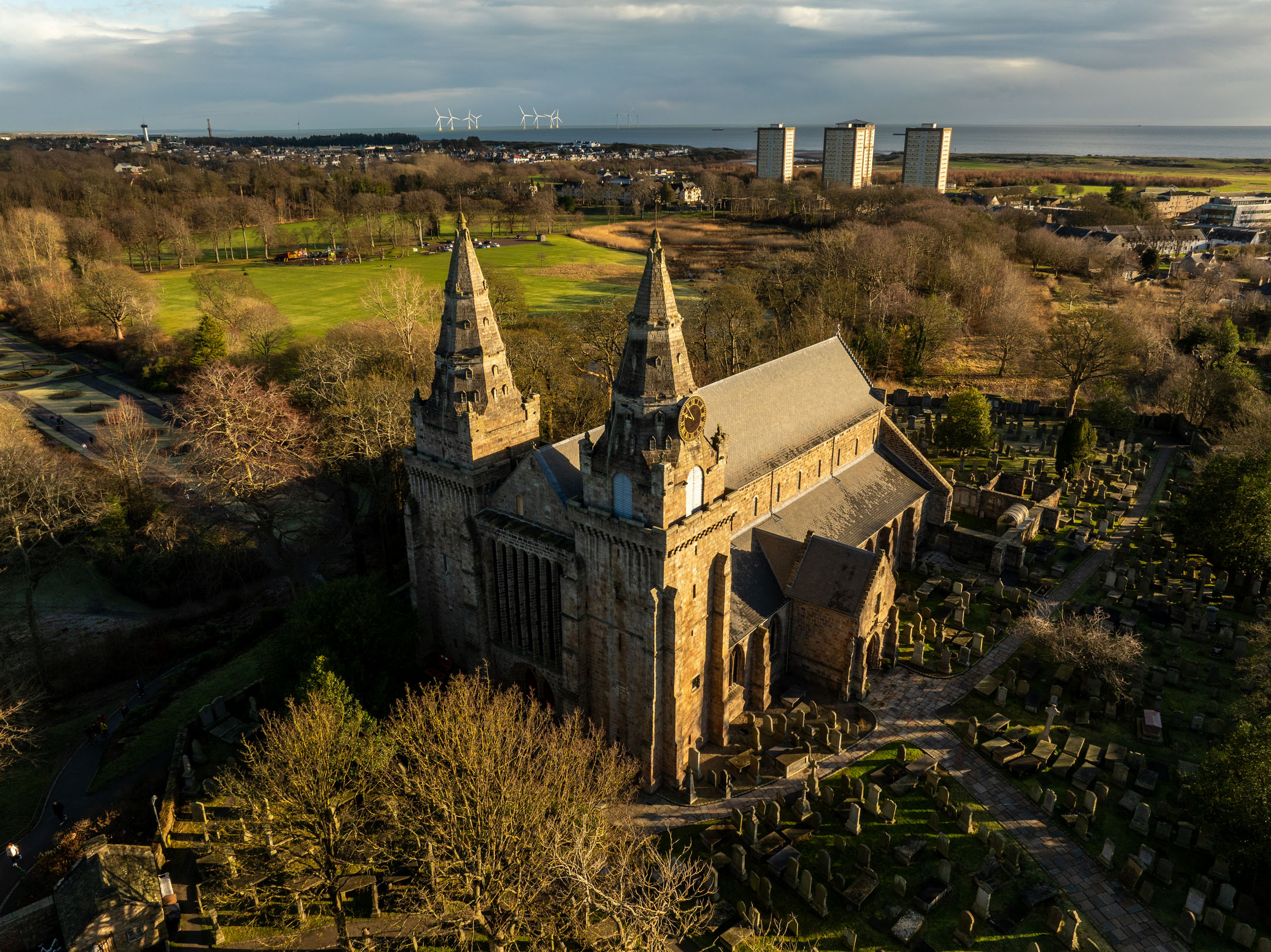 Aerial view of cathedral with twin spires, graveyard and nearby parkland under golden evening light.