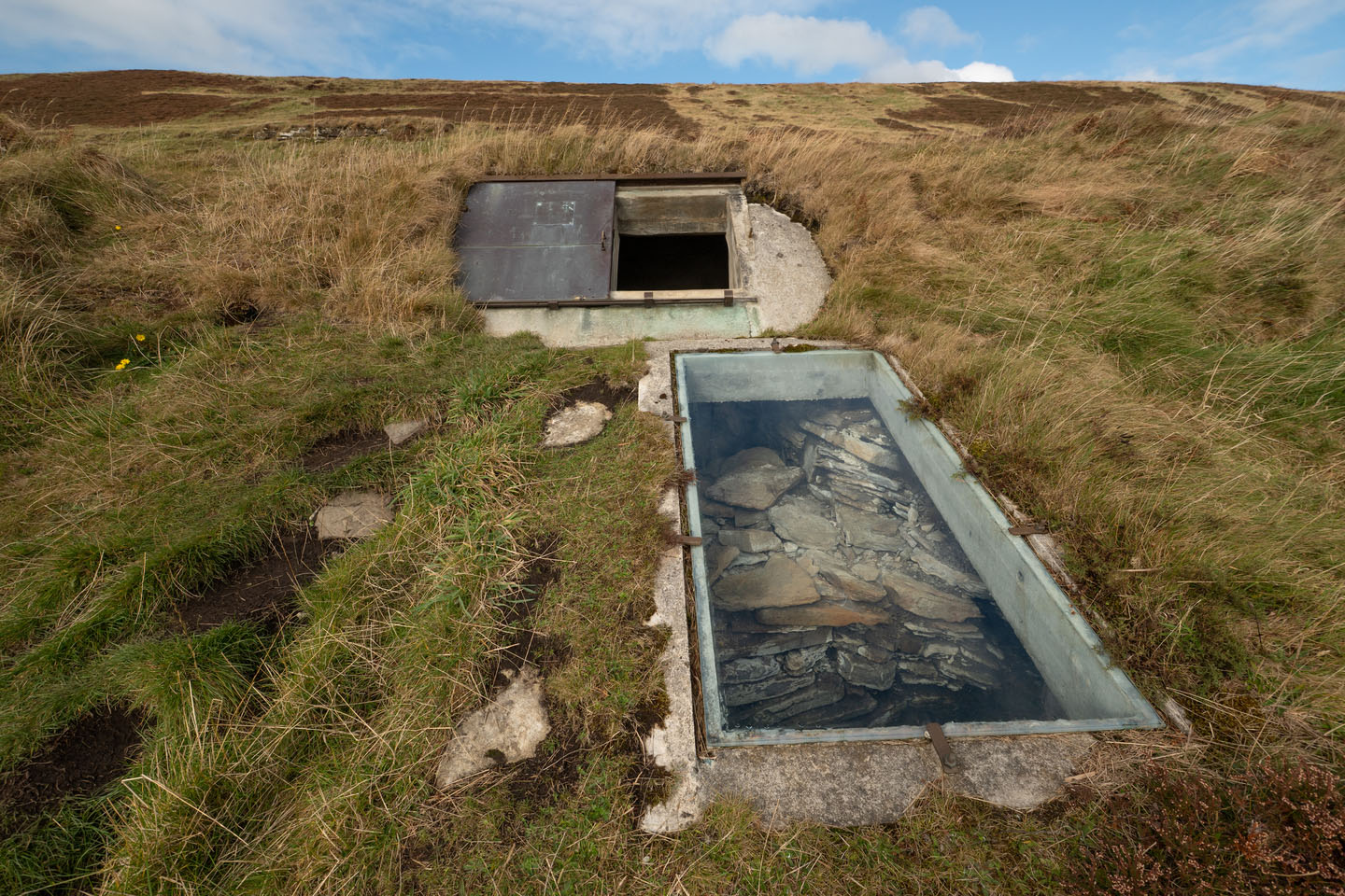 Exterior hatch and glazed rooflight revealing stone chamber beneath the turf mound.