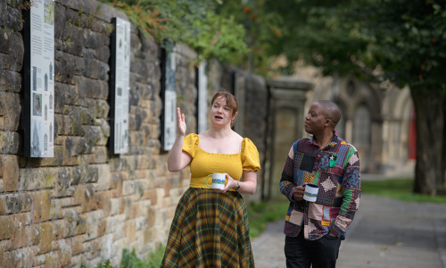 Two people walk alongside an old stone wall talking and holding white mugs with the Talking about Heritage logo on them. In the background out of focus, there is what appears to be an old church.