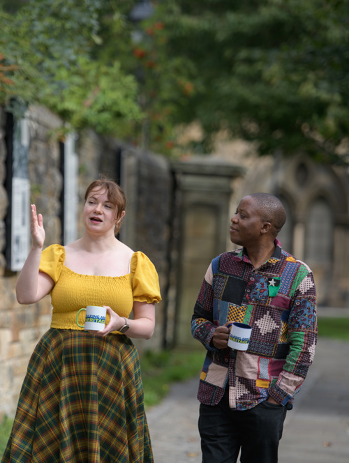 Two people walk alongside an old stone wall talking and holding white mugs with the Talking about Heritage logo on them. In the background out of focus, there is what appears to be an old church.