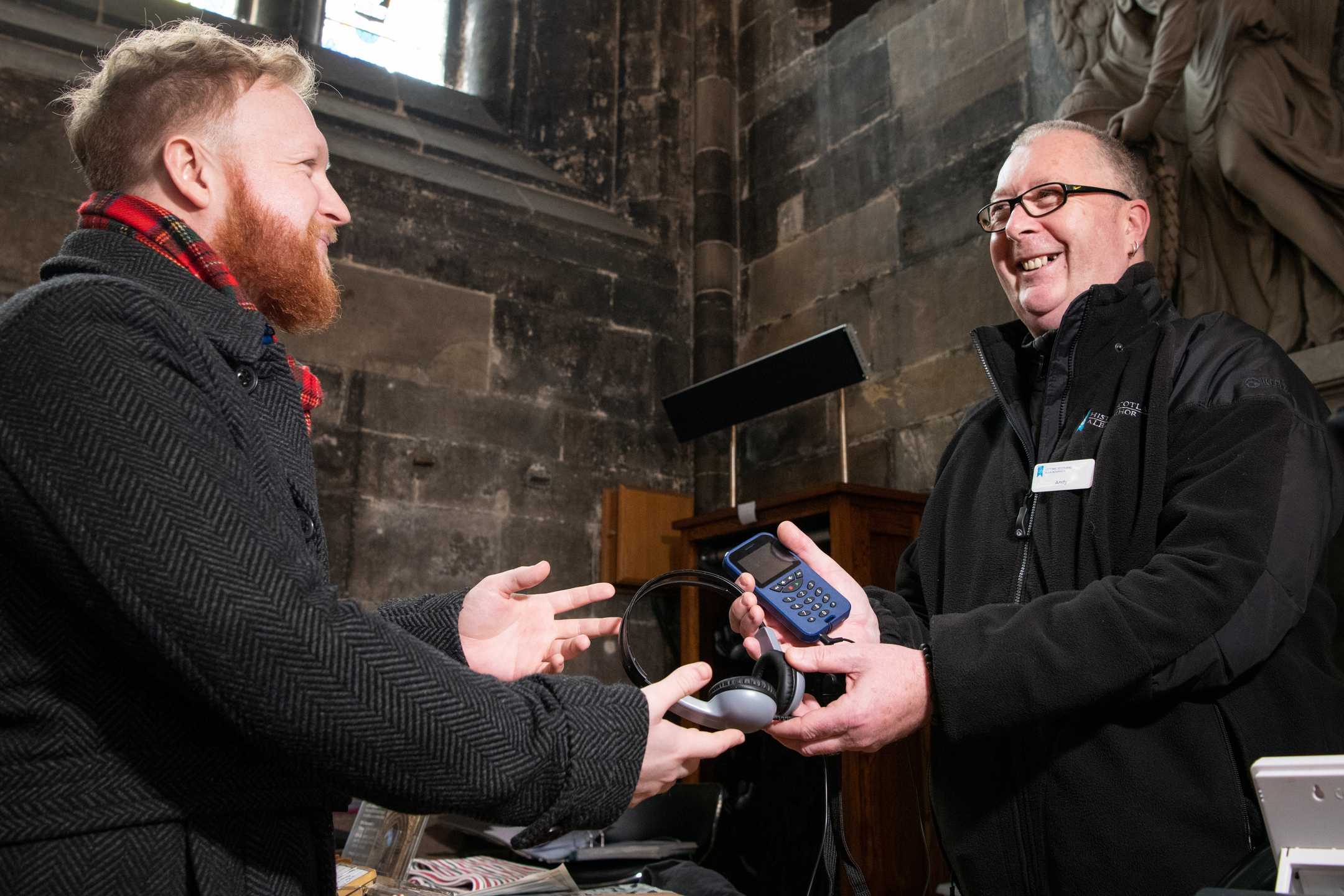 A man in a smart coat and a tartan scarf is handed an audio device and headphones by a staff member at Glasgow Cathedral
