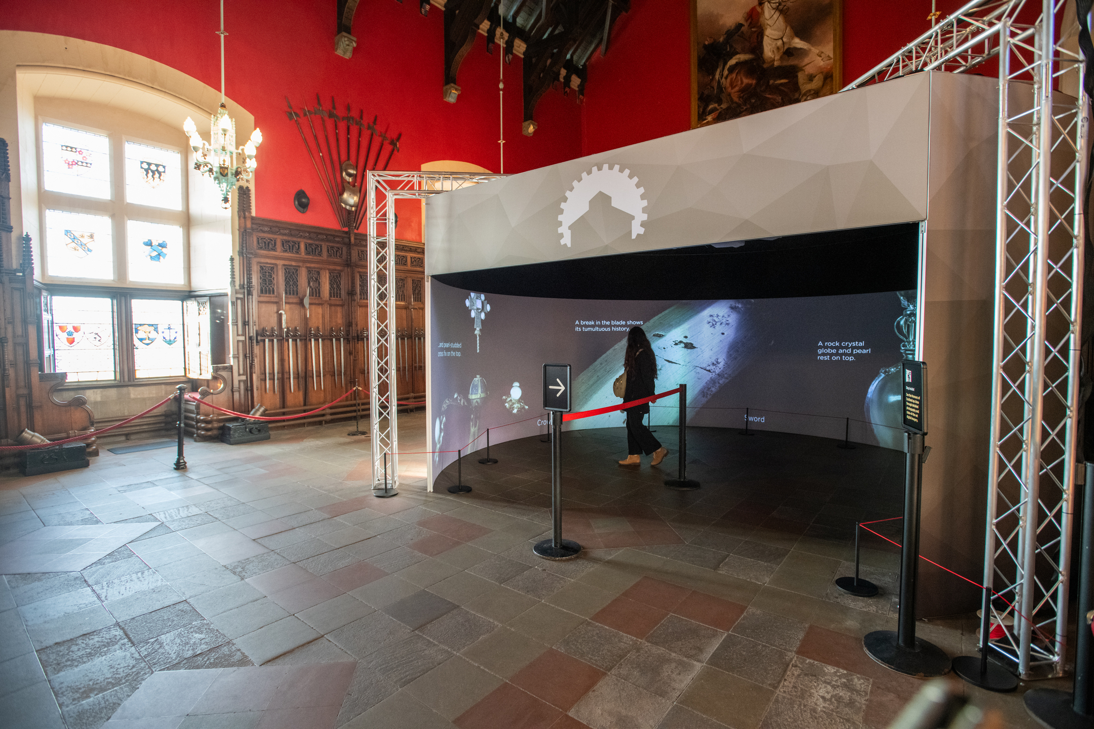 An immersive digital exhibition is installed within a historic hall featuring red walls, stained-glass windows, chandeliers, and displays of ceremonial weapons. A curved projection screen supported by a metal framework shows images and information about historic artefacts, while a visitor walks through the enclosed experience. The contrast between the modern interactive display and the traditional heritage setting highlights the use of digital technology to improve access to Scotland’s historic treasures.