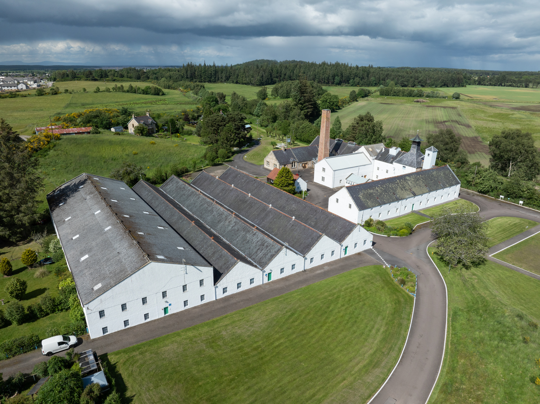 Aerial view of the historic Dallas Dhu distillery complex, showing white warehouse buildings, a tall brick chimney, and surrounding farmland beneath dramatic storm clouds.