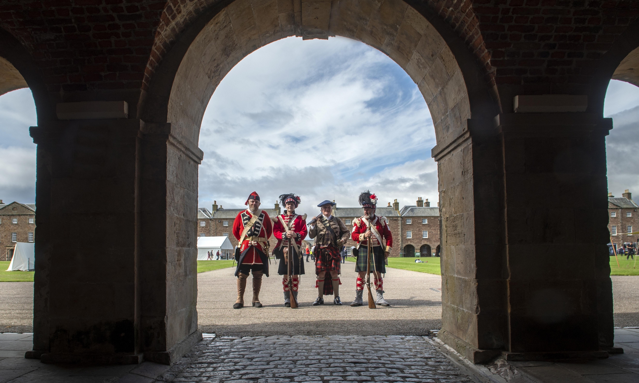 Four historical reenactors in 18th‑century military uniforms stand in the courtyard of Fort George, viewed through a large stone archway.