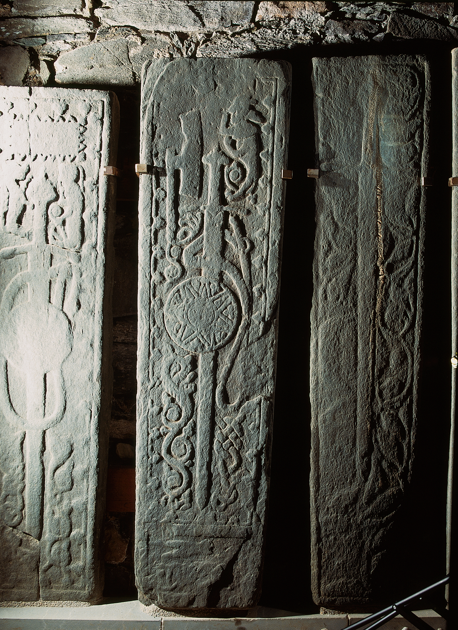 A close-up of two medieval grave slabs displayed upright against a stone wall. Each is carved from dark grey stone and decorated with elaborate motifs—one featuring a long sword with interlaced patterns around it, the other displaying looping vinework and geometric knot designs. The slabs are lit dramatically from one side, casting shadows that highlight the depth of the carvings. The supporting wooden brackets are visible beneath them.