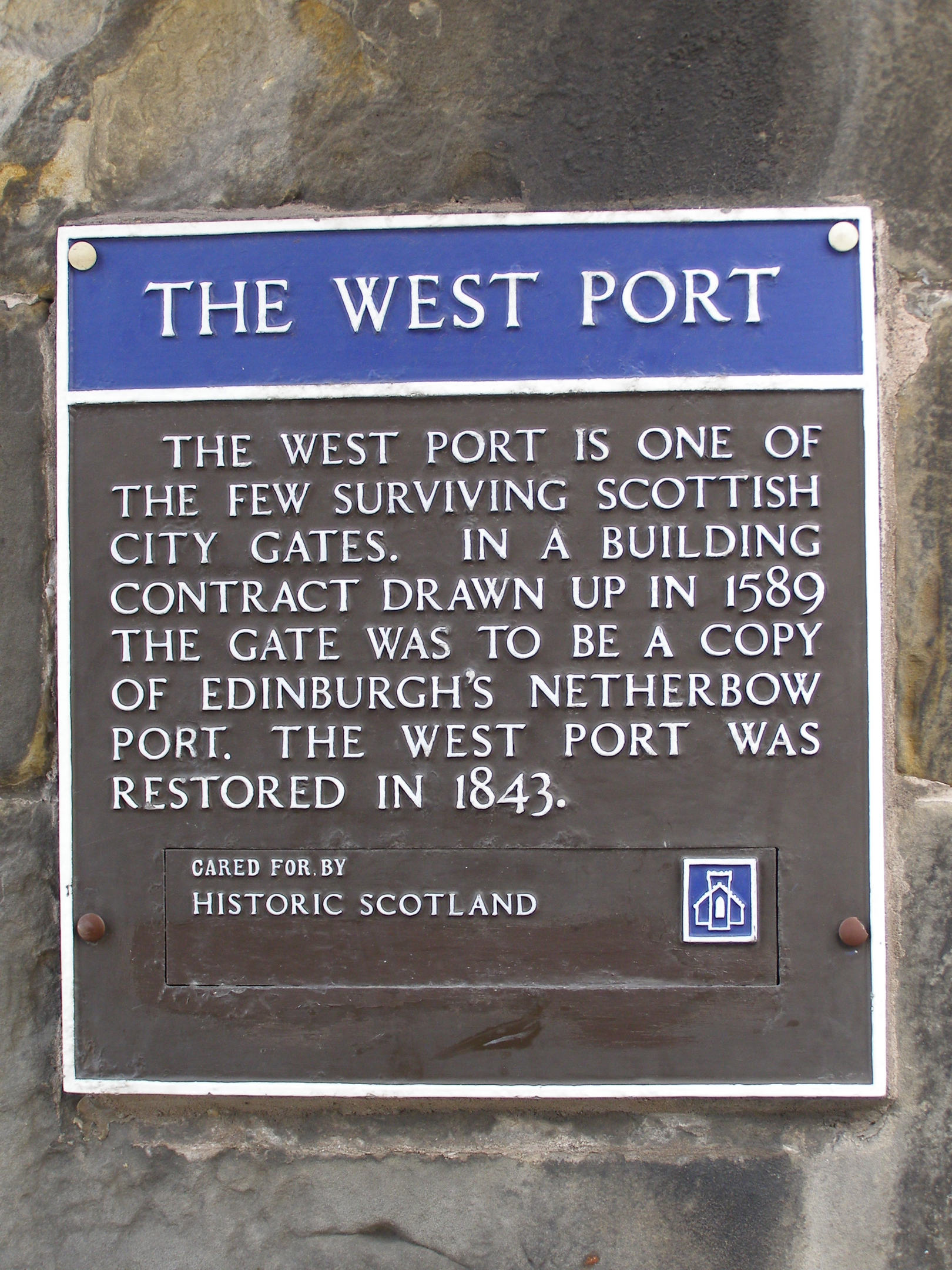 Close-up of a rectangular interpretation plaque mounted on a rough stone wall. The top section has a blue header reading ‘THE WEST PORT.’ Below, engraved white lettering explains the history of the West Port city gate, noting its 1589 design and its 1843 restoration. The bottom of the plaque includes the Historic Scotland logo.”