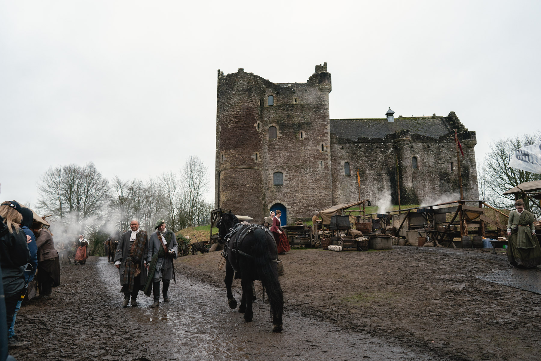 Filming taking place at Doune Castle. Two actors and black horse walk past the castle on a muddy pathway.