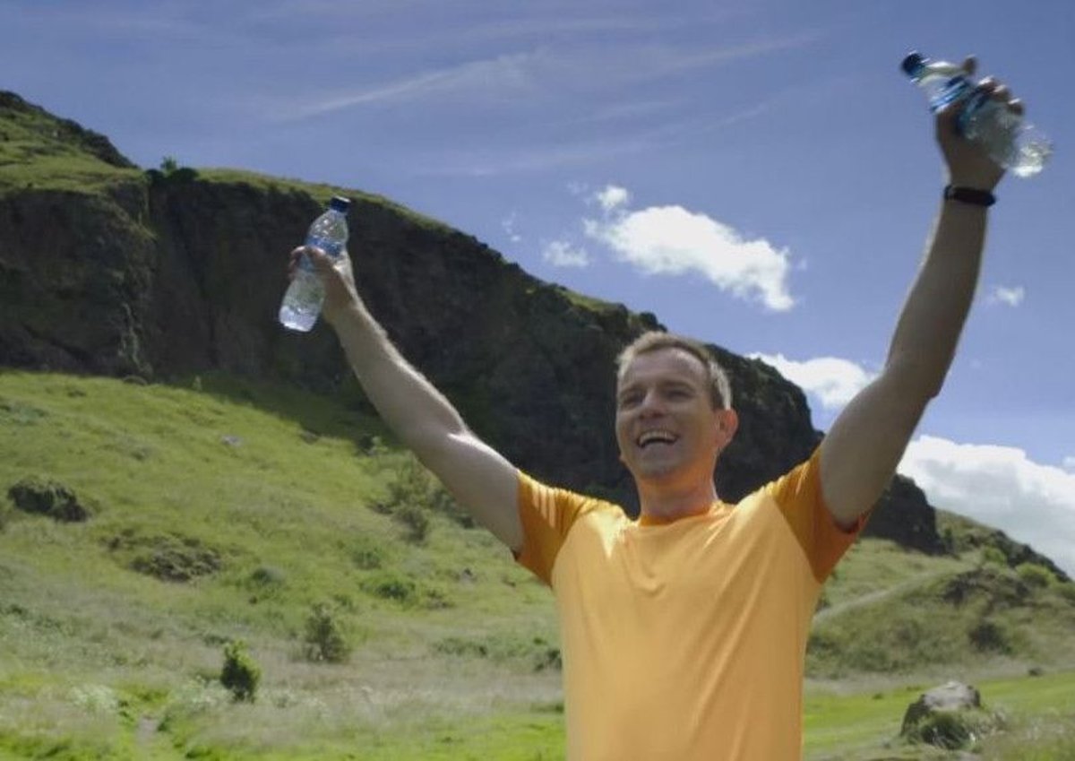A smiling man in an orange T‑shirt raises two water bottles triumphantly while standing in a green valley beneath a rocky hill and blue sky.