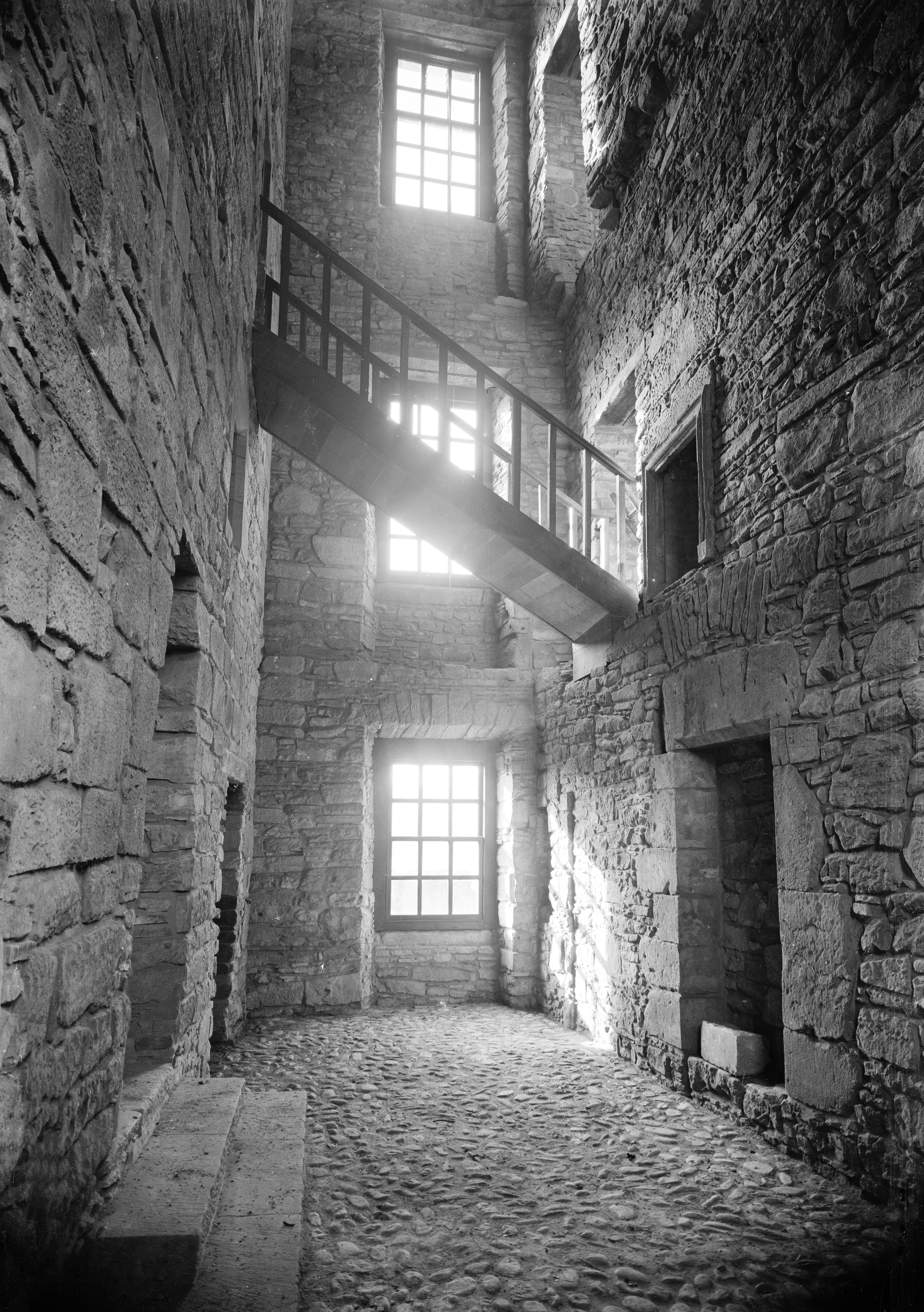 Black and white interior photo of corridor area with stairs crossing and sun shining in through three large windows 