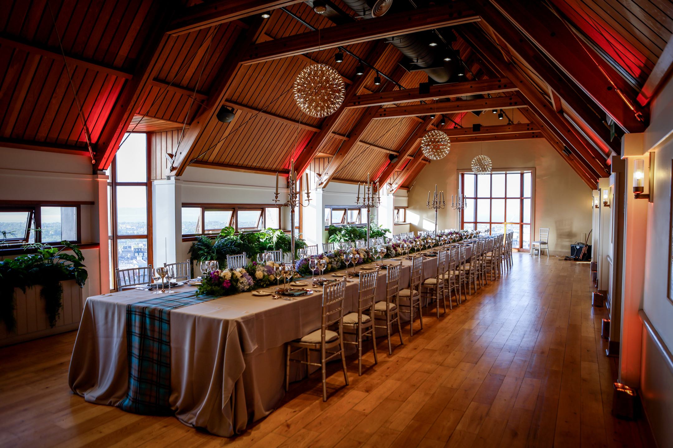 A long banquet table set with tableware, floral arrangements, and tartan table runners inside a bright timber‑beamed hall with a high vaulted wooden ceiling, pendant chandeliers, large windows, and polished wooden floors.