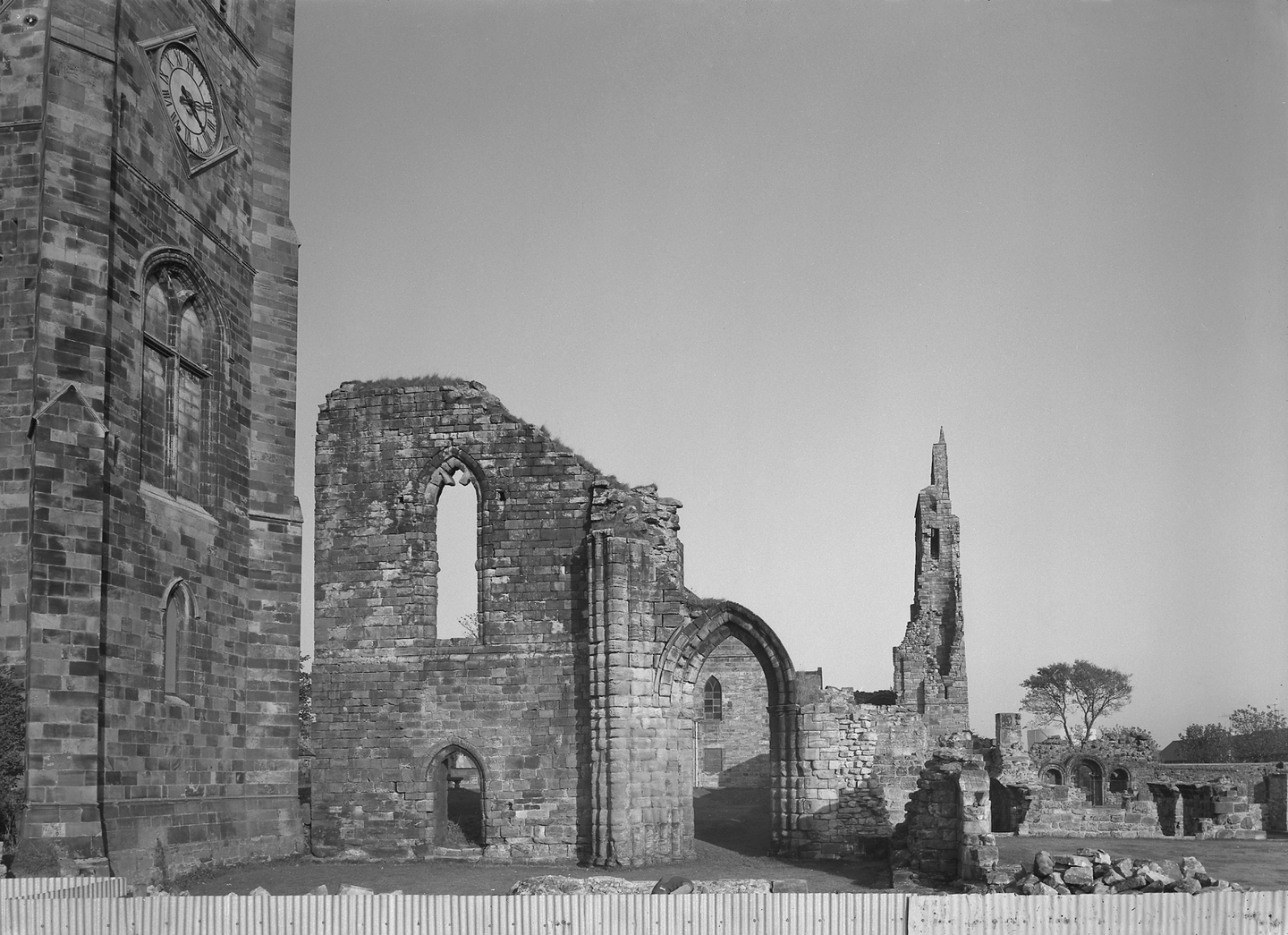 Black‑and‑white photo of a ruined stone church: a tall clock tower on the left and broken walls, arches, and a narrow spire remnant to the right, all under a clear sky.