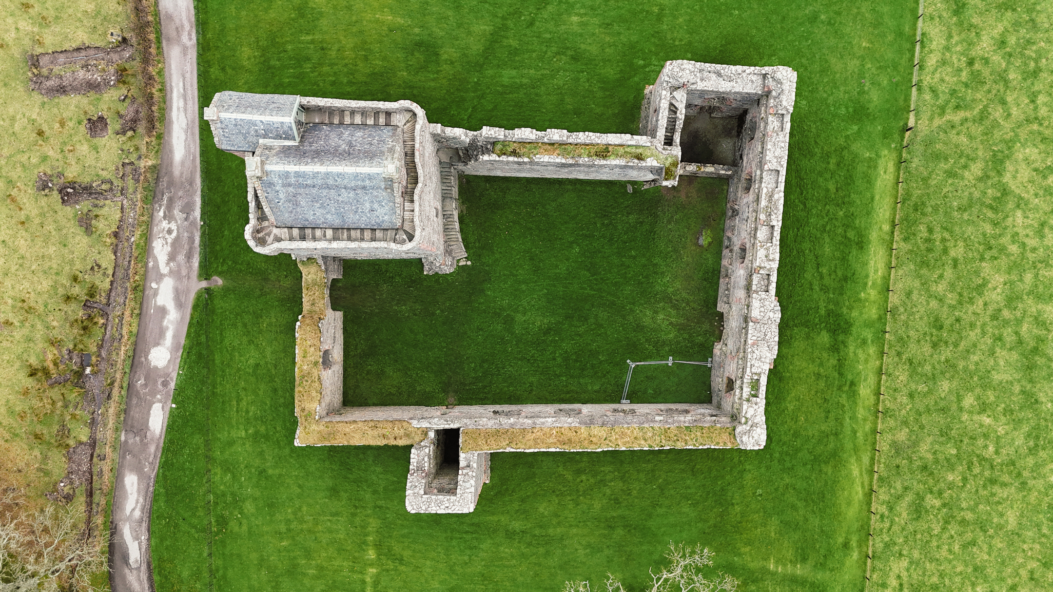 A directly overhead drone shot showing the full outline of the castle’s stone walls, tower, and grassy interior.