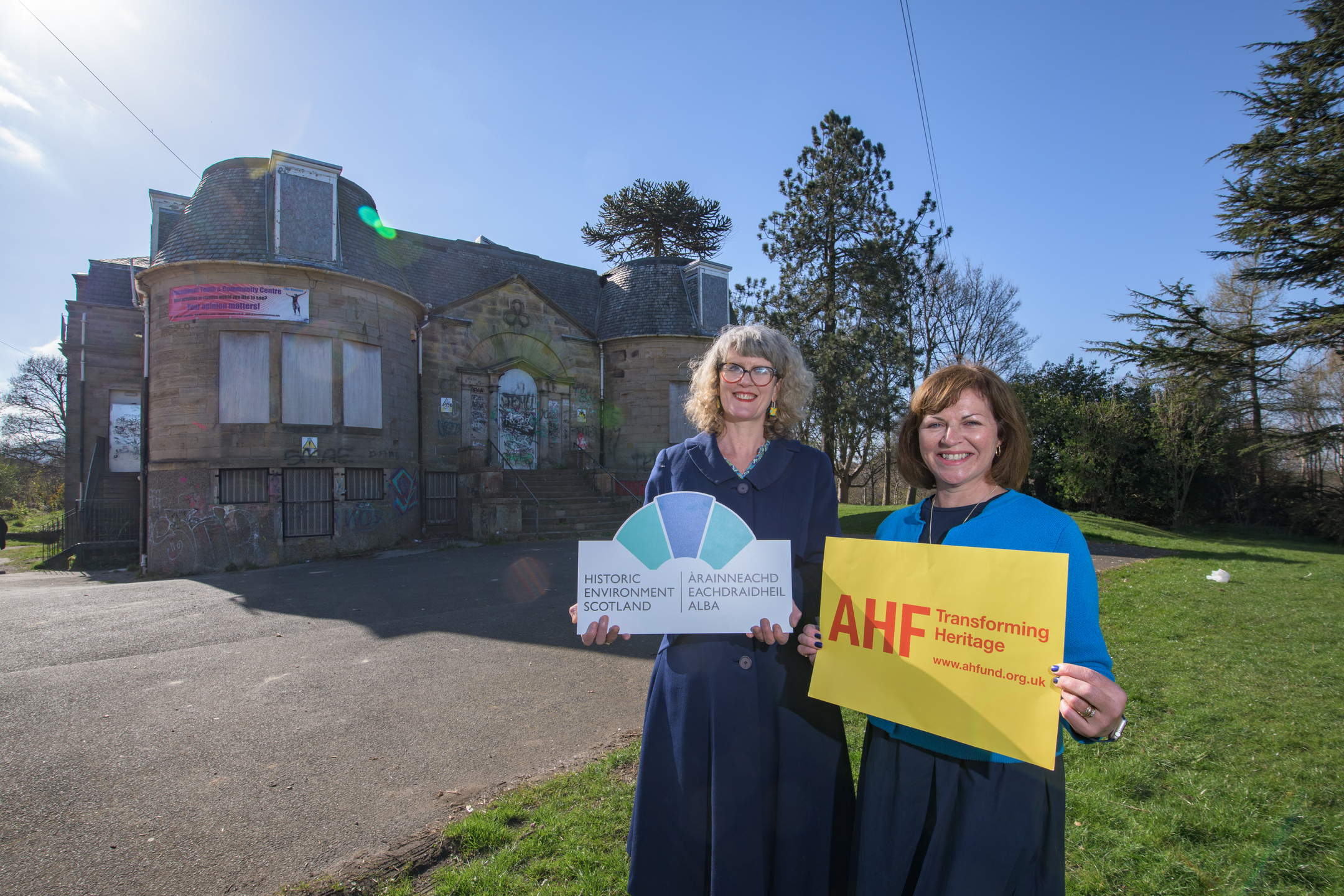 Two women stand outside an old building smiling at the camera and holding up signs, one saying Historic Environment Scotland and the other AHF 