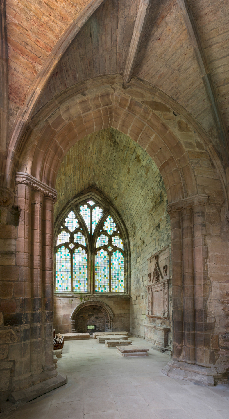 Part of a building, with high ceilings and stone walls. There is a large stained glass window at the end, letting light through. It resembles a church.