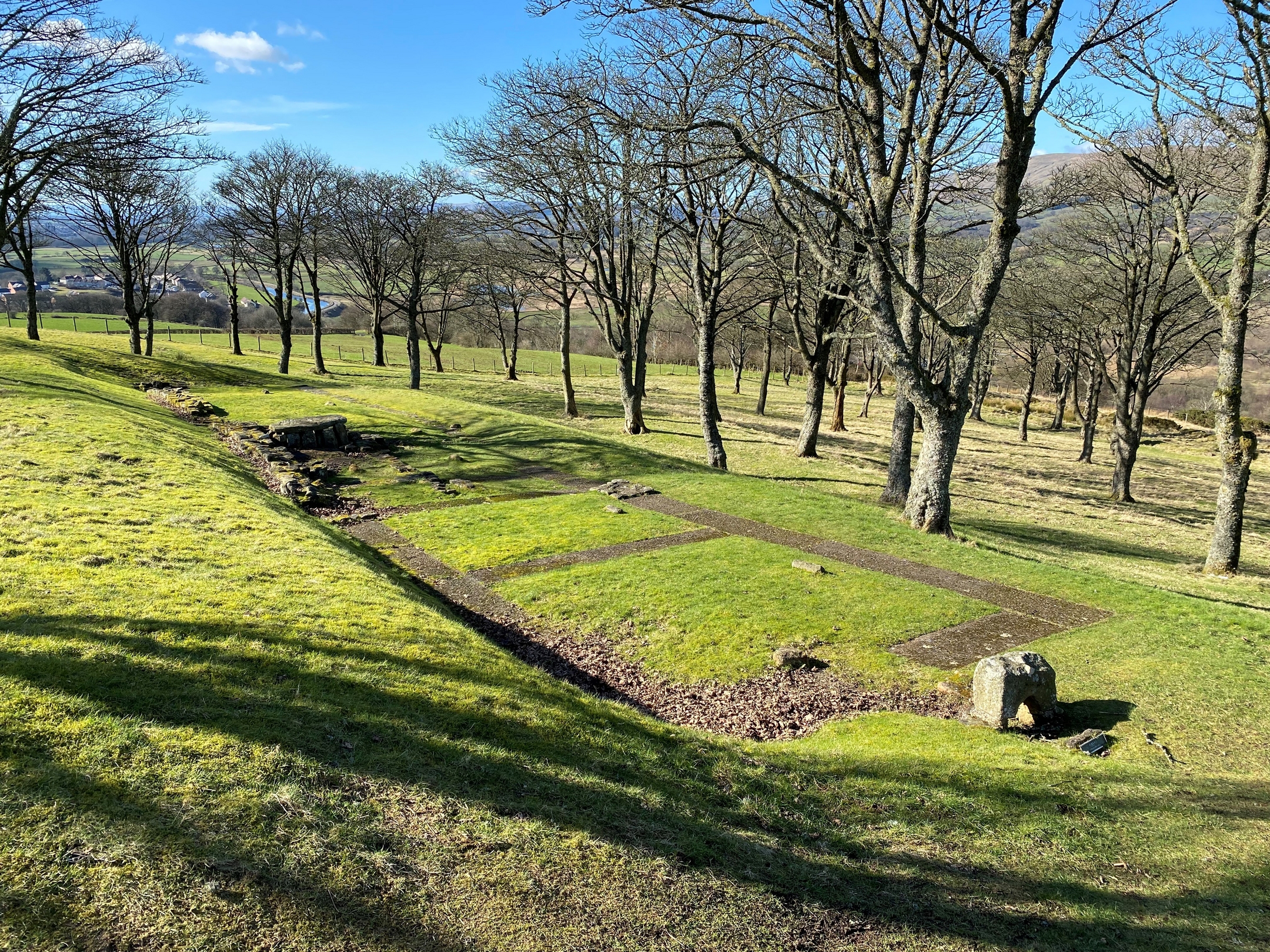 Rectangular stone foundations of a Roman building at Bar Hill Fort, set within a grassy clearing surrounded by leafless trees. The remains outline the structure’s walls, and a stone feature is visible near one end. The landscape slopes gently downward, with distant hills and a village visible through the trees under a bright blue sky.