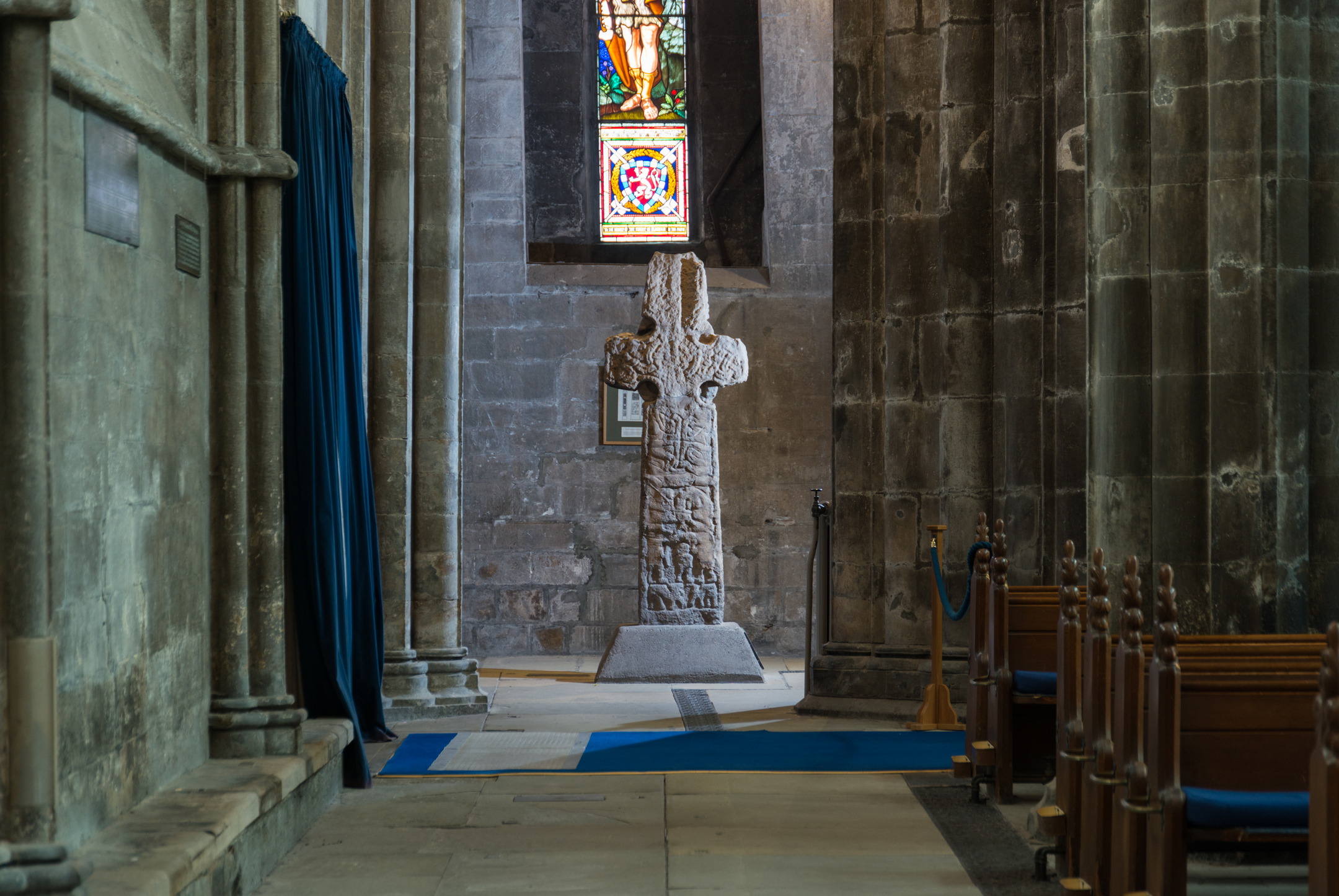 View along the side interior of an abbey with outside wall on the left and pews on the right and a large stone cross at the end of the aisle and stained glass window behind