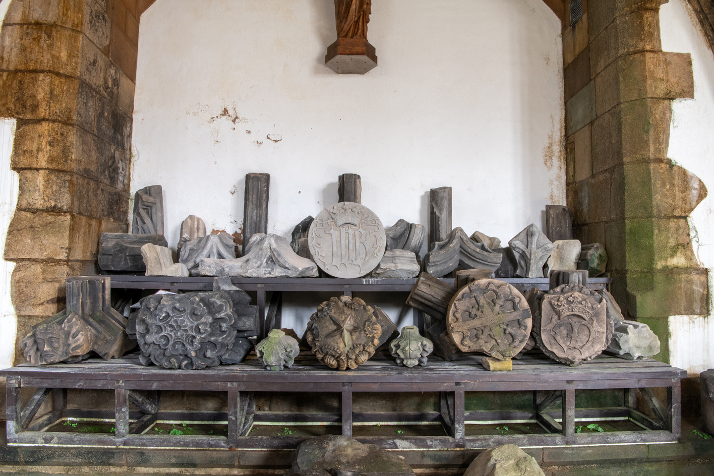 A collection of stone artefacts, some in tact and others crumbled. They sit on a wooden platform of two levels, inbetween an stone brick arch. Some of the stones in a better condition have markings on them.