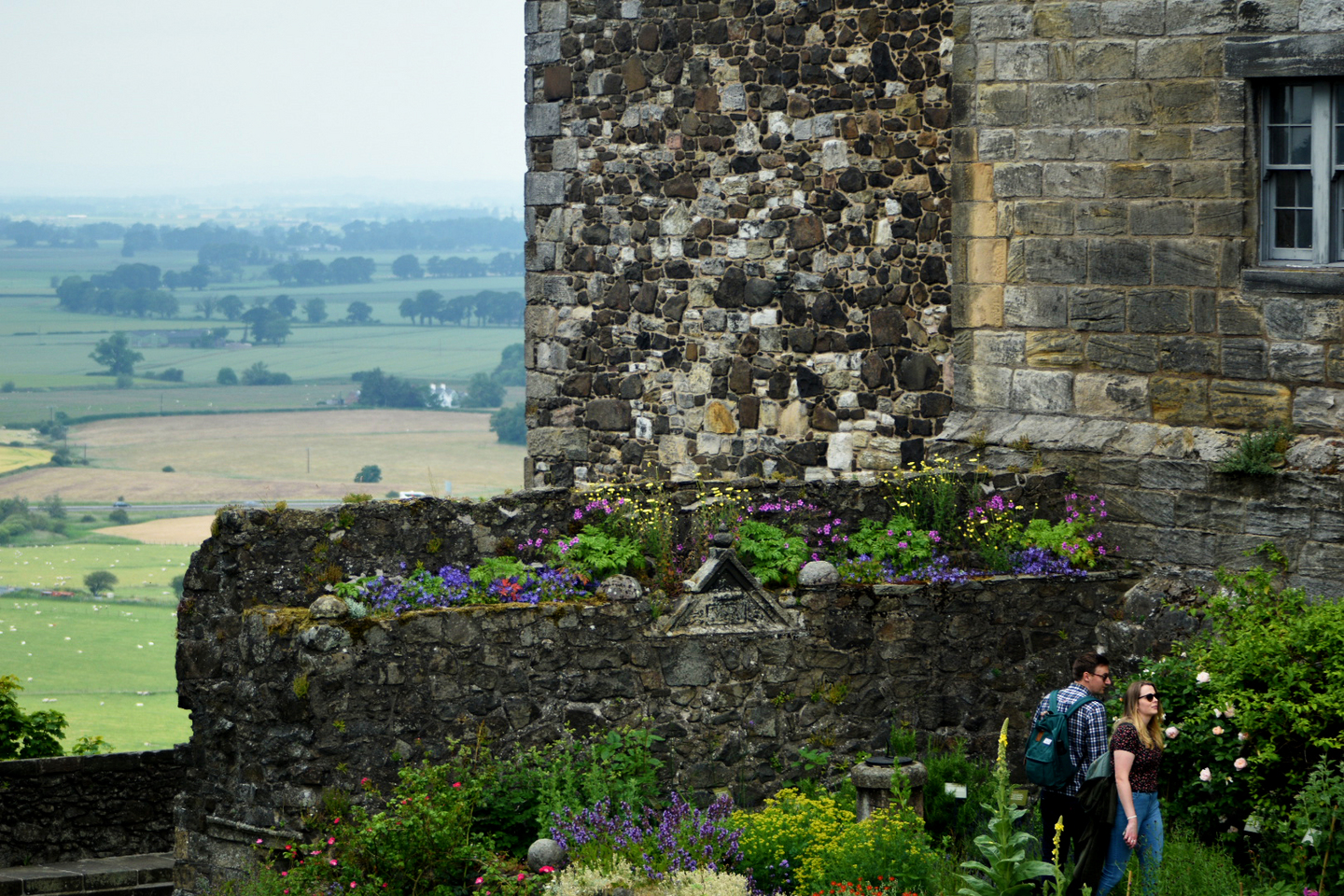 Side of a stone building, with a stone platform next to it where flowers are growing. There are flowers growing in front of the platform, with two people exploring the gardens. There is a rural view to the side of the building. 