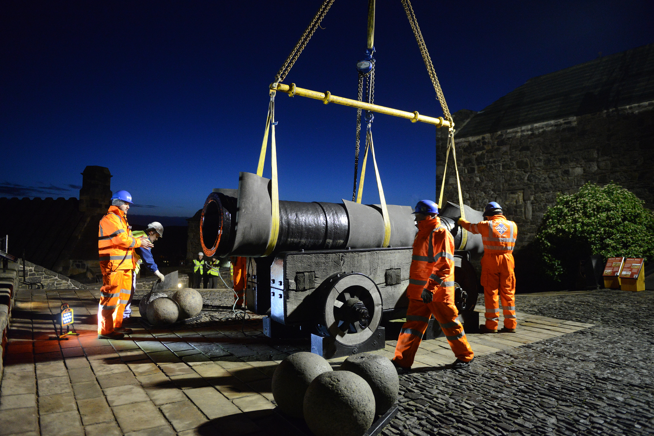 A team in orange hi-vis work on securing Mons Meg, a large cannon, onto a crane strap
