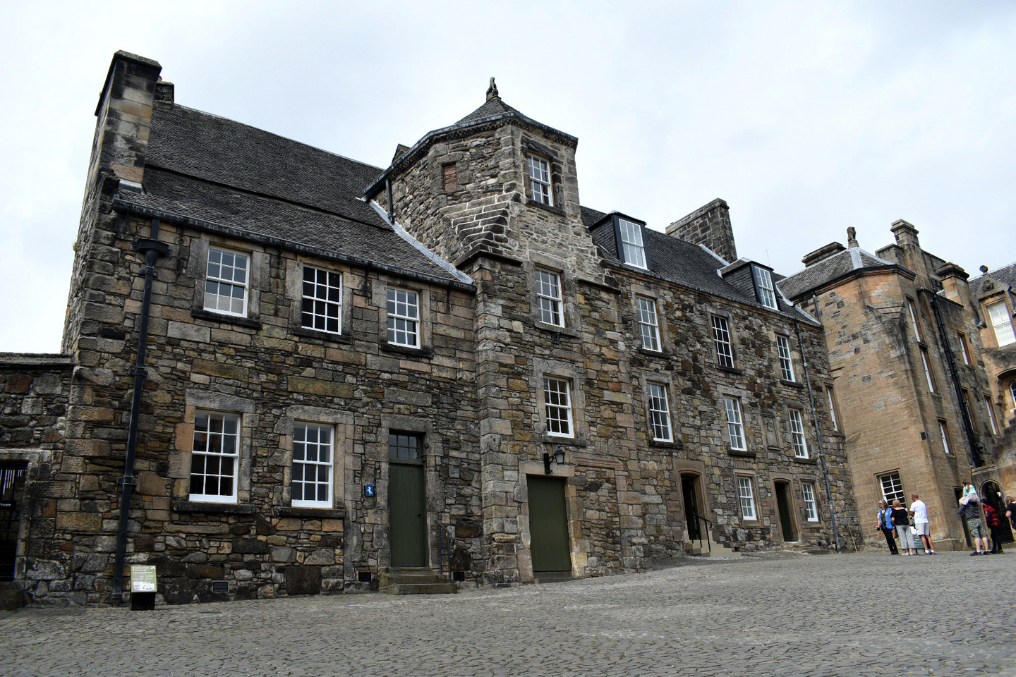 Exterior view of a large historical building made of stone. There are multiple windows and doors of varying sizes on it. Some of the stone has changed colour with age. There are people standing observing the building. 