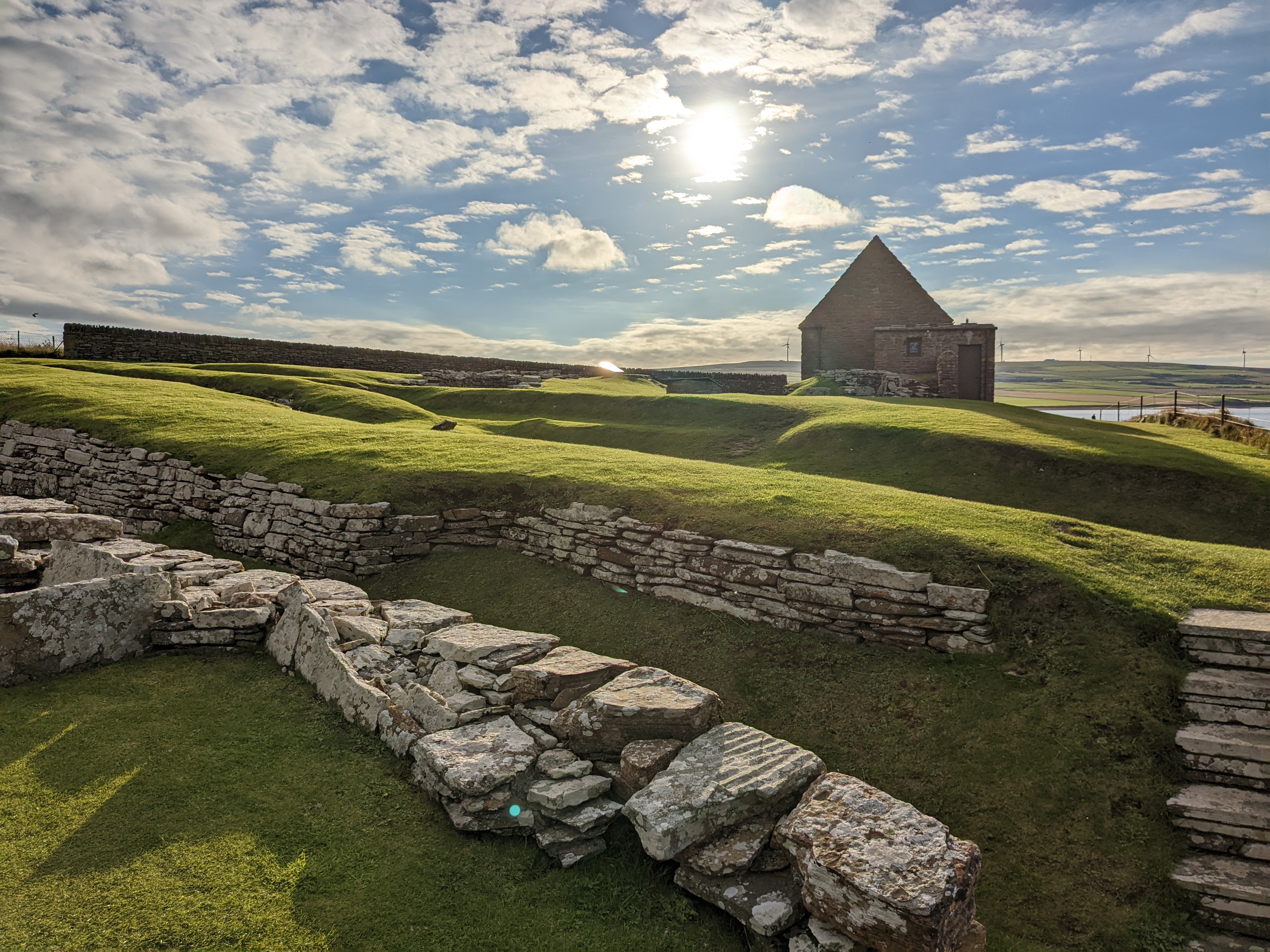 Grassy mounds and stone walls of the Broch of Gurness under bright sunlight, with a small stone-roofed building in the background. The sky is filled with scattered clouds and the sea is visible beyond.