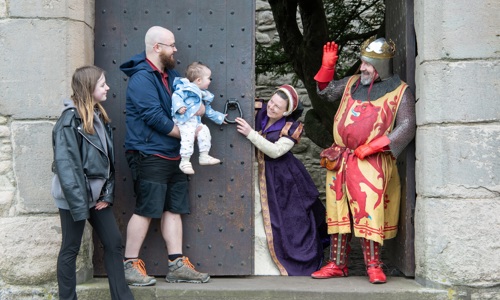 A teenage girl and adult man who holds a baby girl stand outside the large wooden door of a castle. They are greeted by a woman in a purple dress and a man in armour wearing a crown