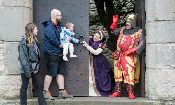 A teenage girl and adult man who holds a baby girl stand outside the large wooden door of a castle. They are greeted by a woman in a purple dress and a man in armour wearing a crown