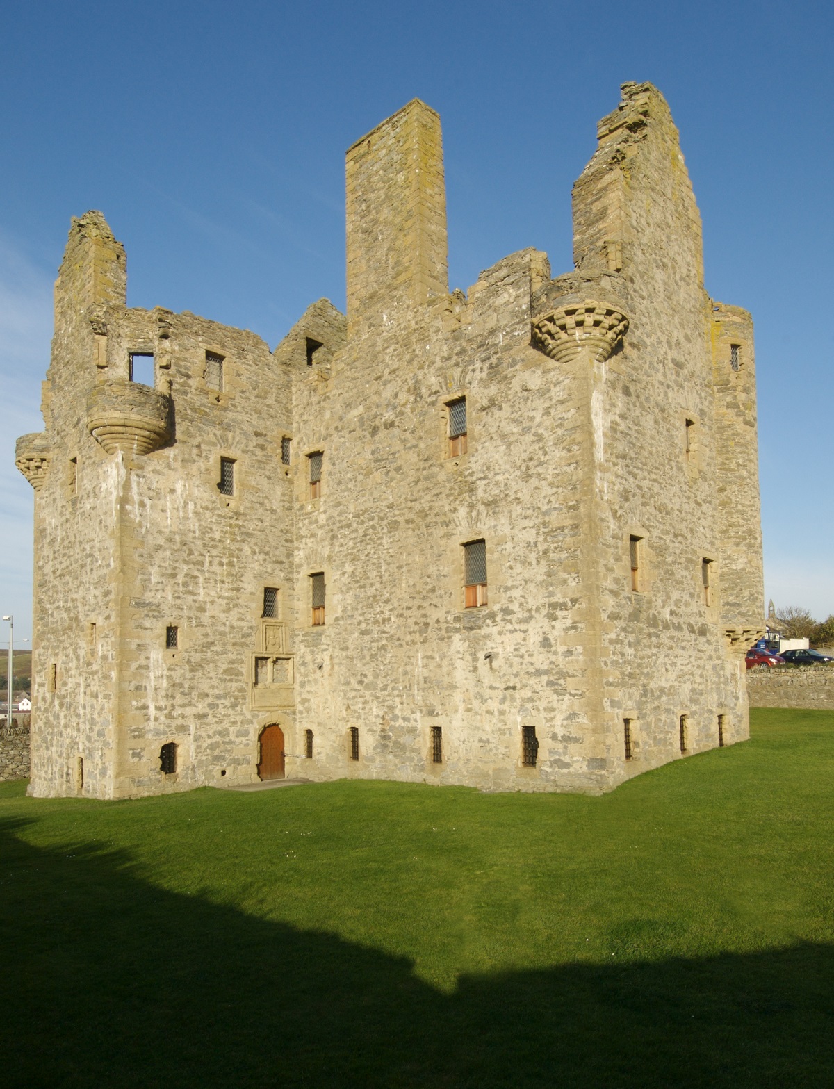 Exterior view of Scalloway Castle, showing its tall, roofless stone walls, corner turrets and narrow windows, standing on a grassy lawn under a bright blue sky.