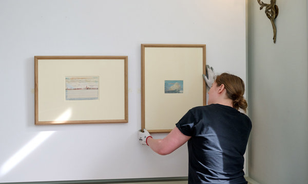 A woman wearing white gloves adjusts a mall watercolour painting in a wooden frame on a white wall, next to another watercolour painting. Both artworks depict landscapes.