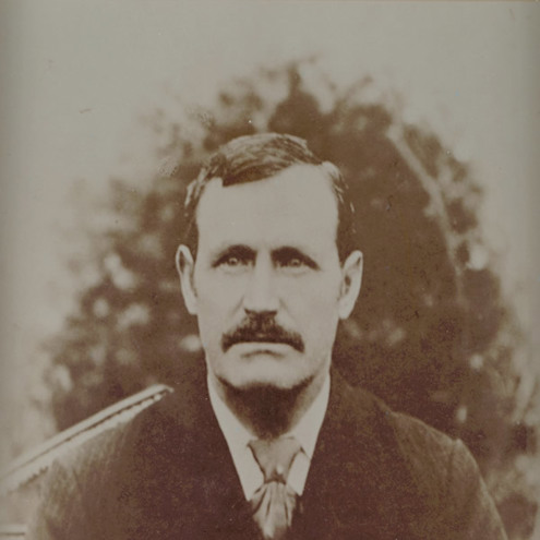 Sepia toned, front on portrait photo of gentleman in a suit, with a large moustache and short black hair parted on the side.