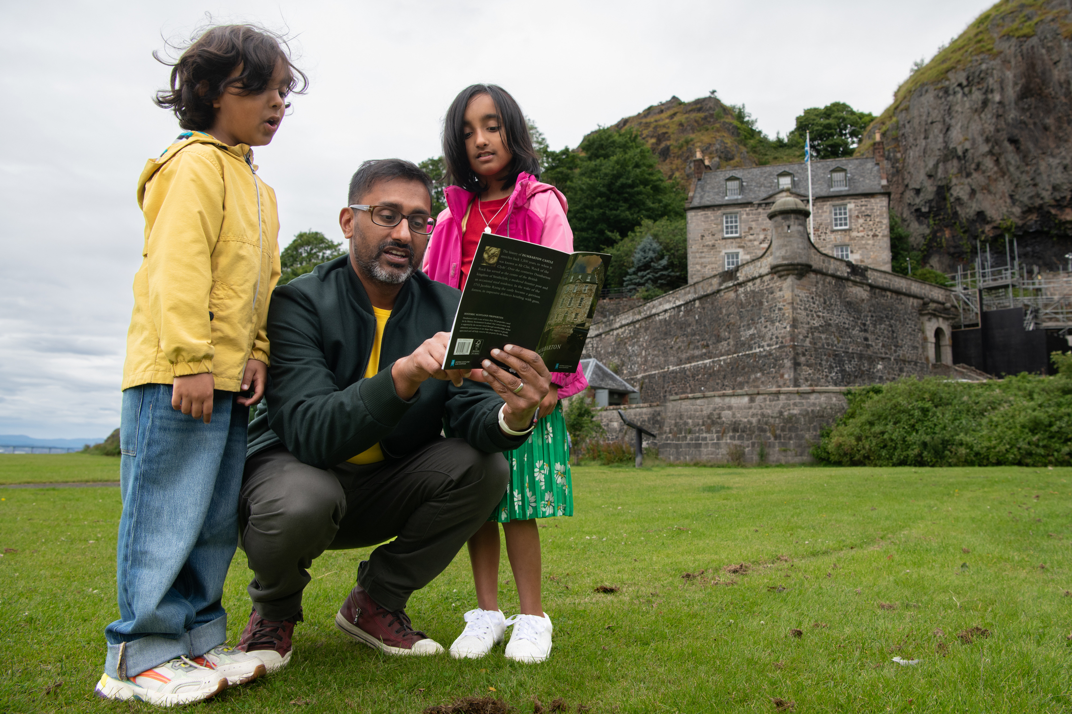 An adult kneels on the grass with two children while reading a guidebook, with stone castle buildings in the background.