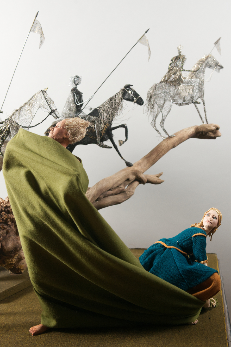 A model of a cloaked figure in a green mantle kneels beside a smaller figure in blue, both looking toward a backdrop showing wire‑framed mounted soldiers with lances. A large piece of driftwood forms part of the scene’s landscape.