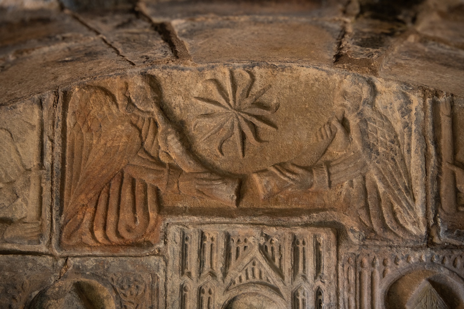Close-up of a stone carving showing two winged figures holding a circular, star‑like symbol between them.