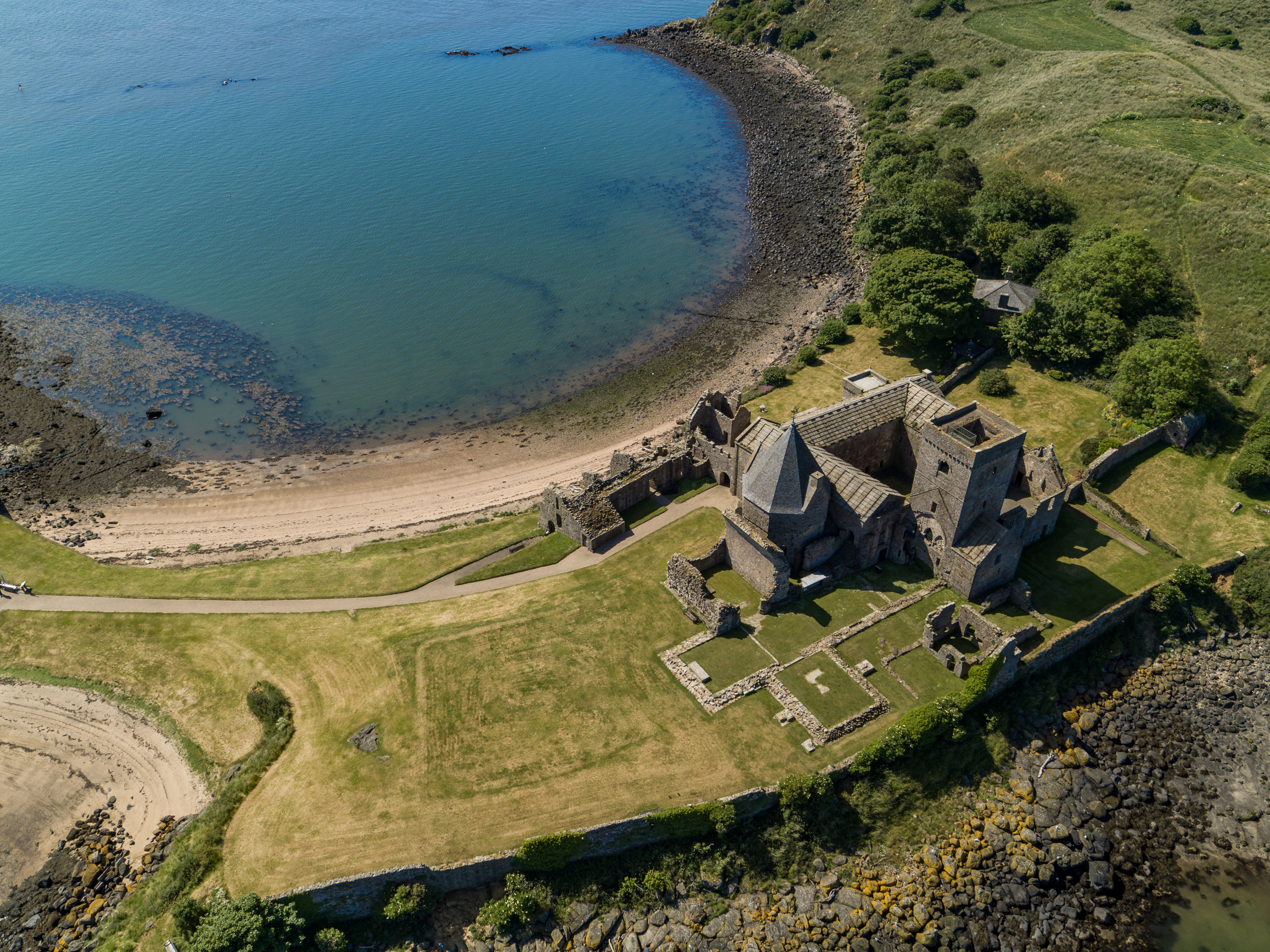 A large, stone historic abbey complex on a grassy island, beside a sandy beach