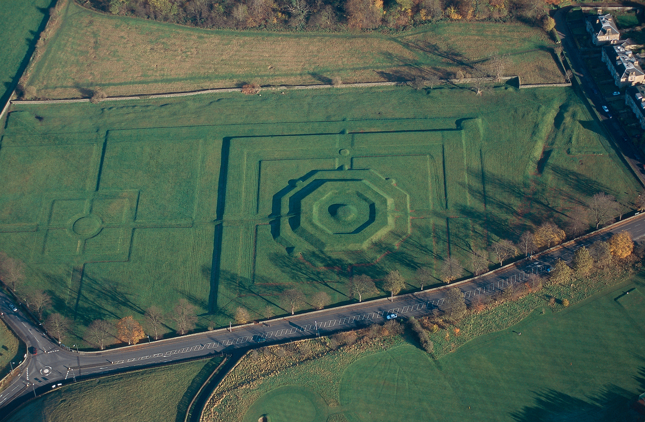 Aerial view of a large, geometric earthwork set in green parkland, featuring an octagonal raised mound at the centre, straight embanked paths, and formal rectangular outlines beside a nearby road and houses.