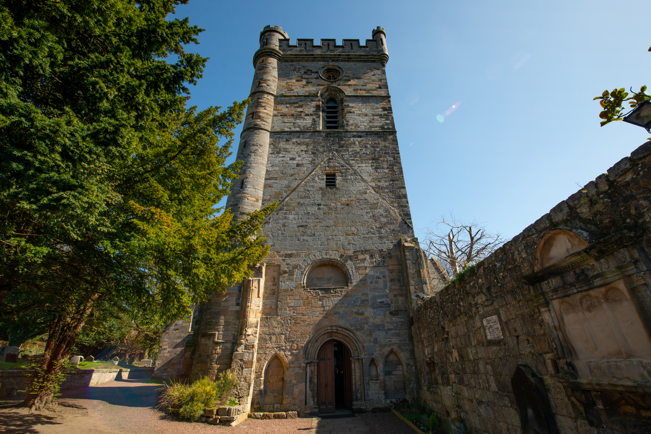 A tall view of Culross Abbey’s tower rising above the entrance doorway, with trees lining the gravel path and sunlight casting long shadows.