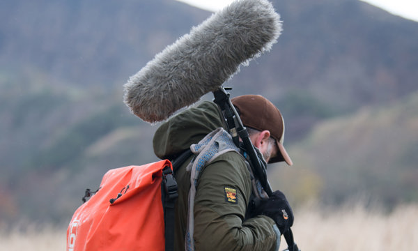 A man wearing a baseball cap and orange backpack carrying a boom microphone over his shoulder