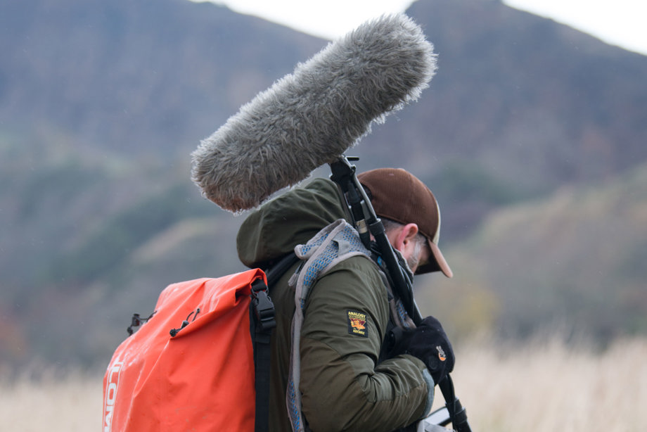 A man wearing a baseball cap and orange backpack carrying a boom microphone over his shoulder