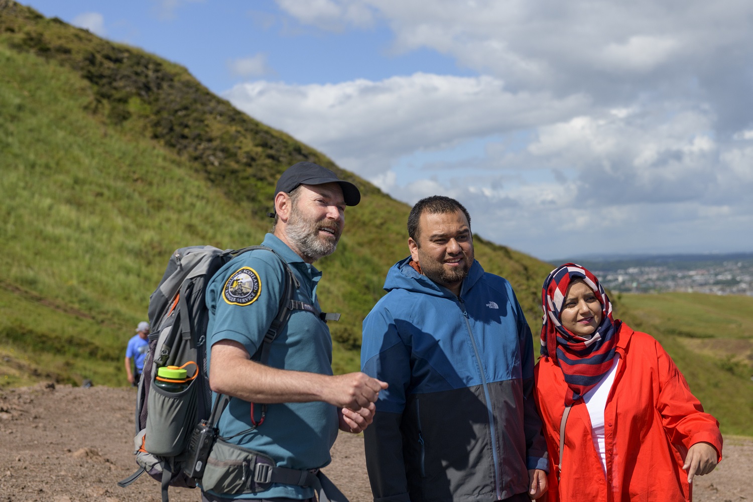 A ranger wearing outdoor gear and carrying a backpack stands on a hillside path talking with two visitors. Green slopes rise behind them under a bright, partly cloudy sky, with views of the city in the distance.