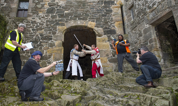 Two re-enactors in the grounds of a castle battling with swords and a crew of 4 with cameras, videos and clapper board